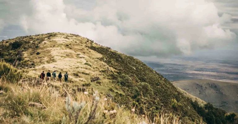 A small group of hikers walks up a grassy highland ridge under heavy cloud, with wide plains far below