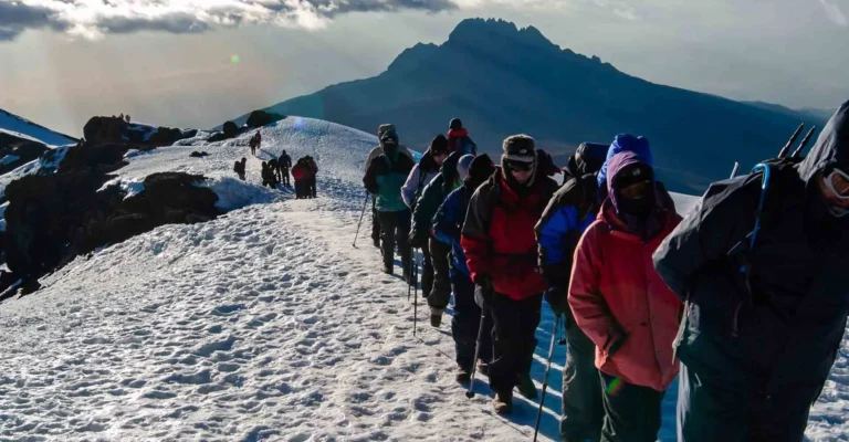 A line of climbers in heavy jackets walks a snowy path near Kilimanjaro's summit, with Mawenzi peak beyond