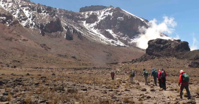 A small group of trekkers crosses a wide, dry alpine plain below Kibo's glacier-streaked cliffs and drifting cloud