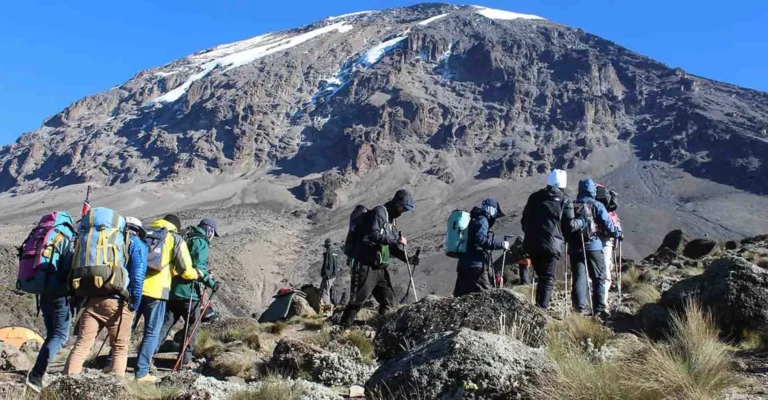 A group of trekkers with colourful rucksacks heads up a rocky trail towards Kibo's steep, snow-streaked face