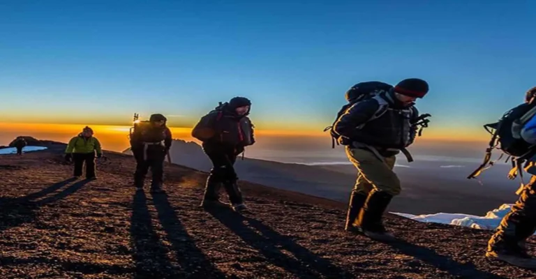 Climbers in down jackets walk along Kilimanjaro's upper slopes at sunrise, long shadows stretching across the volcanic scree