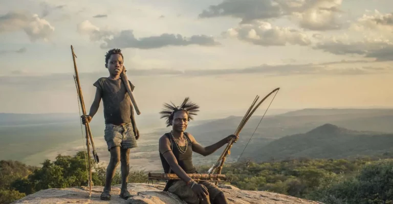 Two Hadzabe hunters holding bows and arrows on a rocky outcrop above a wide hazy valley at dusk