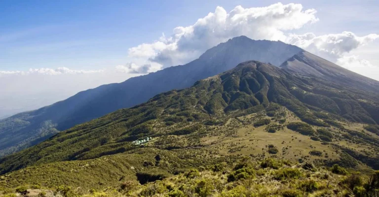 The summit cone of Mount Meru rises above green moorland slopes, with a cluster of huts on a ridge
