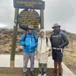 Two trekkers and their guide beside the Kilimanjaro National Park sign at Morum Picnic Site, Shira Plateau, 3,407 m