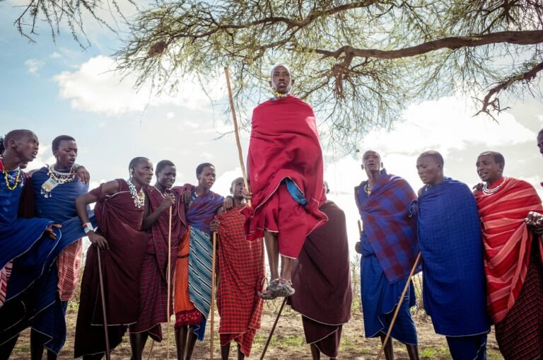 Maasai men and women in red and blue shukas and beaded collars watch as a young man leaps high in the traditional adumu jumping dance under an acacia tree in Tanzania
