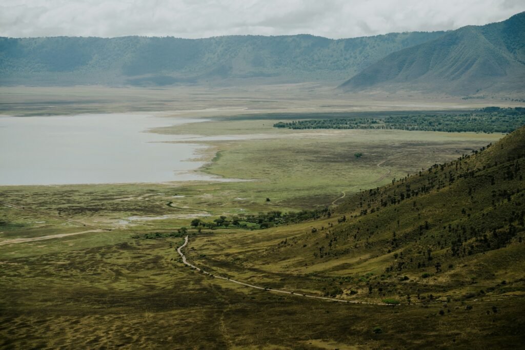 View from the rim down into Ngorongoro Crater: a descent road winds across green slopes toward the crater floor and Lake Magadi, with the far crater wall under cloud