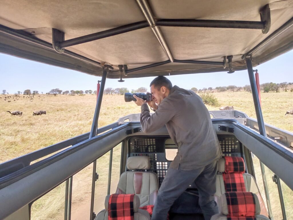 A traveller photographs wildebeest from the open pop-up roof of a safari vehicle on the grassy plains