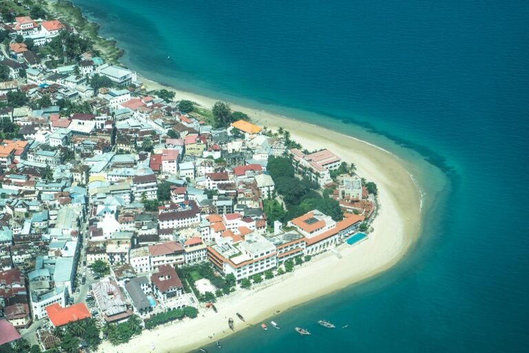 Aerial view of Stone Town, Zanzibar: dense whitewashed and red-roofed buildings along a curving sandy waterfront, with small boats moored in the turquoise Indian Ocean