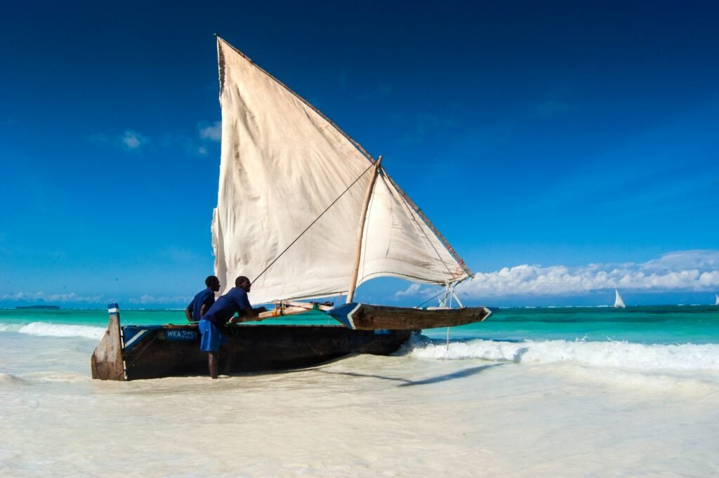 A wooden dhow with a raised cream sail pulled up on the white-sand beach at Matemwe, Zanzibar, turquoise water and a second sailing dhow on the horizon under a deep blue sky