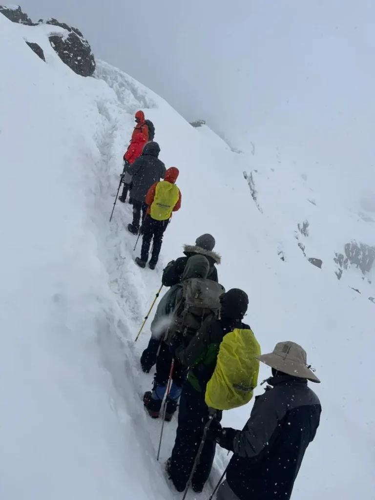 A group of climbers with rain covers on their packs follows a narrow path cut into deep snow as flakes fall