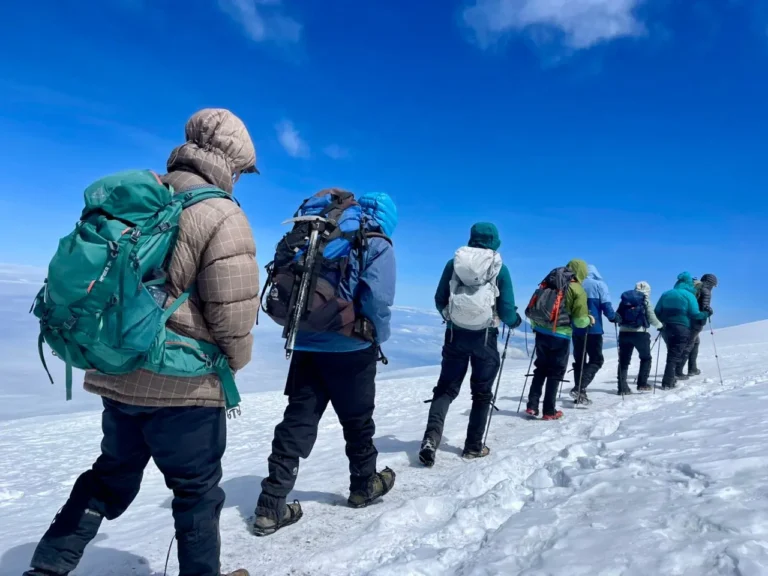 A line of climbers in down jackets and crampons walks across a snowfield high on Kilimanjaro, clouds far below