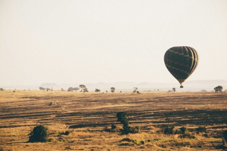 A hot-air balloon drifts low over golden Serengeti grassland dotted with acacia trees in the morning haze